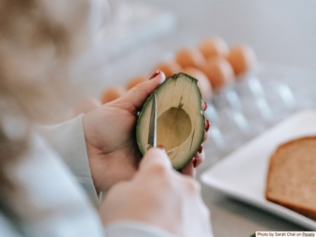 Hands slicing thin brown layer off cut avocado to reveal fresh green flesh underneath on a wooden board