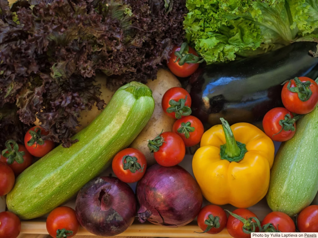 Hands reaching into an open refrigerator produce drawer filled with colorful vegetables including peppers and zucchini