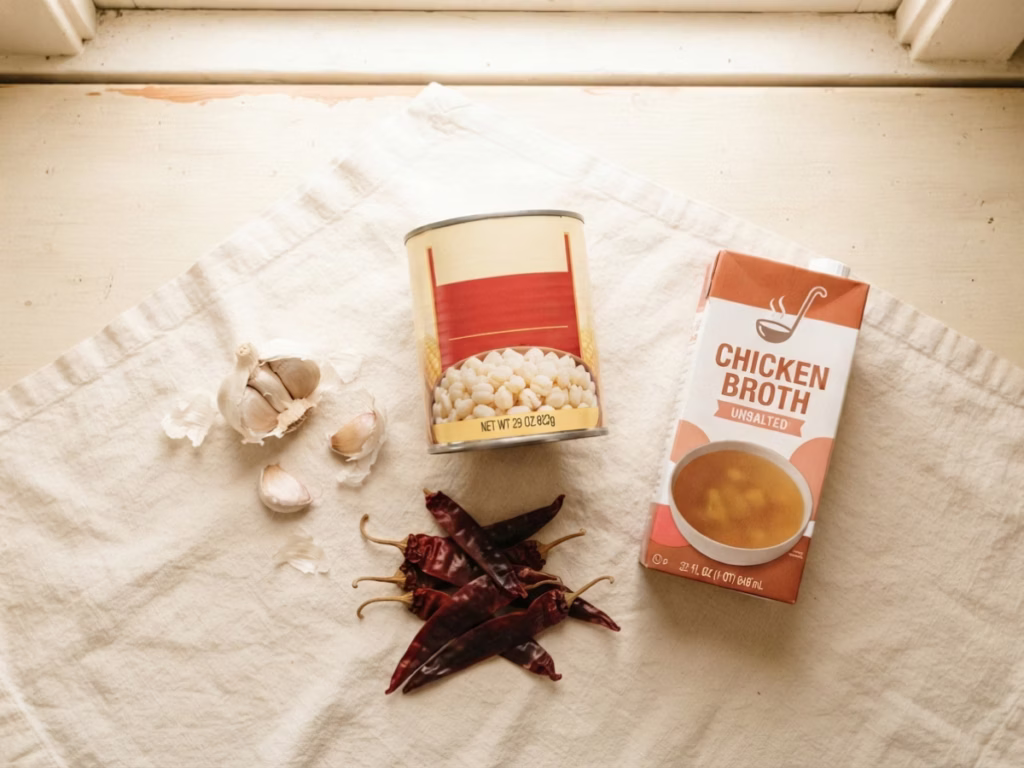 Overhead view of pozole pantry staples: canned hominy, dried chiles, garlic bulb, and broth carton on a cream counter